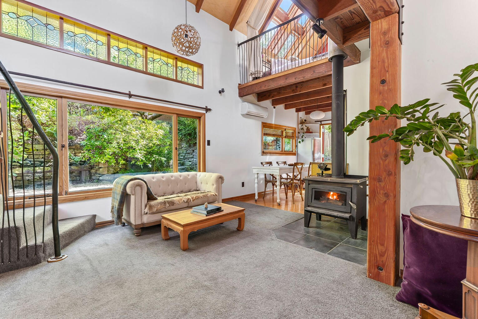 Open-plan living space with large windows, a wood-burning stove, exposed beams, and lush garden views