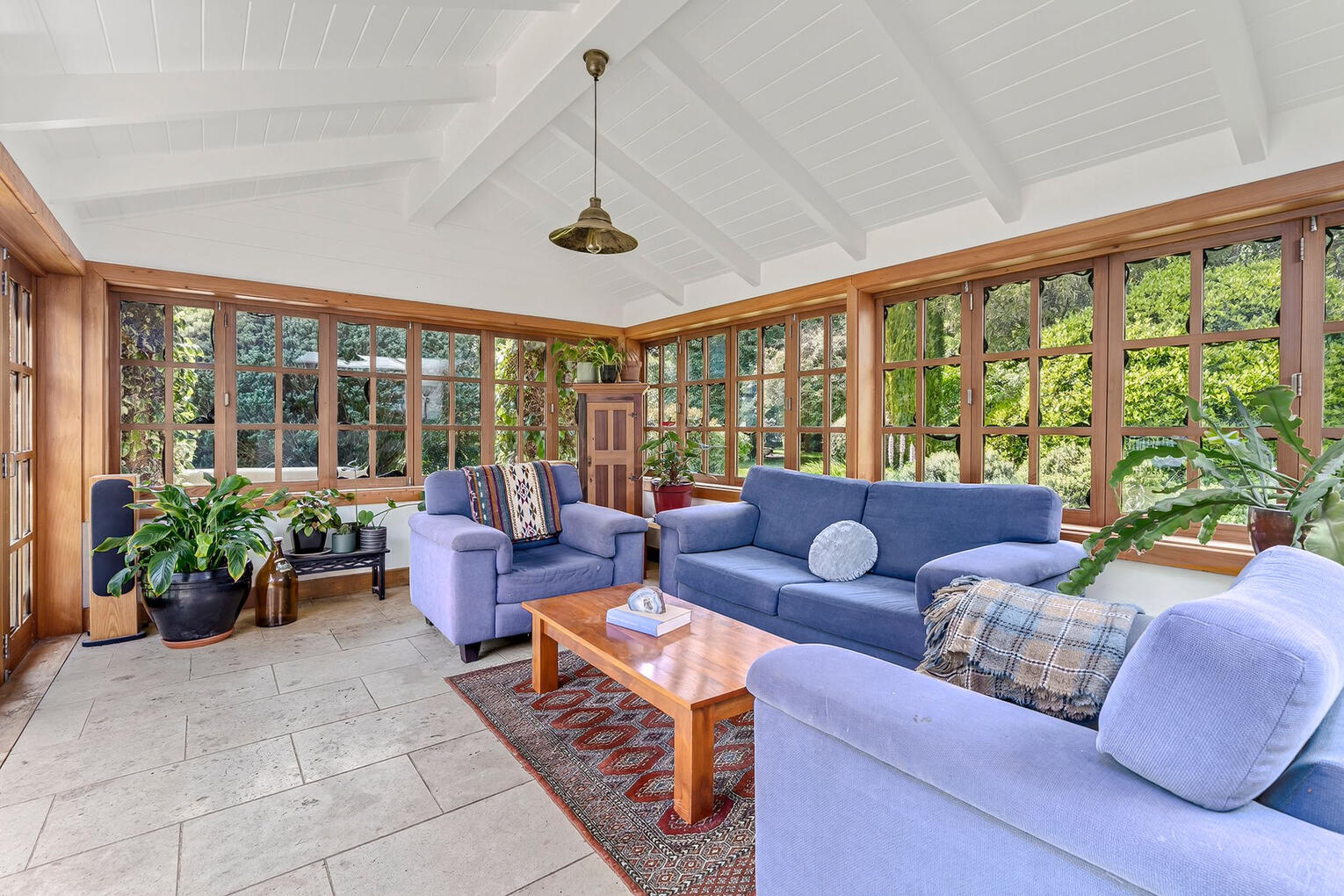 Sunlit conservatory lounge with floor-to-ceiling windows, lush greenery views, and comfortable blue seating